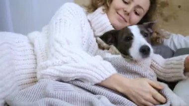Gülümseyen kadın, köpeğin battaniyenin altında beklediği rahat yatağa tırmanır. Sahibi yumuşak yatakta evcil hayvanına nazikçe sarılıyor. Geniş yatakta sıcacık kucaklaşmalar, insan ve hayvan arasında atmosfer rahatlığı yaratıyor.