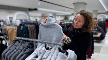 Gri balıkçı yakalı kazağı dikkatle inceleyen bayan alışverişçi moda butiğinin içinde askıda asılı, kışlık kıyafet seçeneklerini araştırıyor ve rahat örgü elbise seçimi yapmayı düşünüyor.