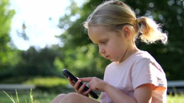 At kuyruklu sarışın kız çimenli parkta oturuyor, akıllı telefon ekranına derinden konsantre oluyor, güneşli yaz gününde tamamen dijital aktivitelere odaklanıyor. Çocuklar ve aletler için kavram