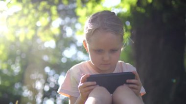 Yapraklı dalların arasından süzülen ılık güneş ışığı, çimenli zeminde oturan genç kız, açık havada huzurlu bir ortamın tadını çıkarırken akıllı telefon ekranına dalmış. Çocuklar ve aletler için kavram