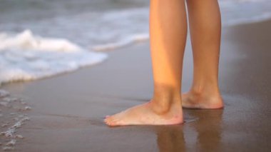 Çıplak ayaklı çocuk bacakları ıslak kumlu sahil şeridinde hareketsiz duruyor. Küçük dalgalar, altın saat güneş ışığı altında minicik ayakları hafifçe yıkıyor. Çocuklarla kumsalda eğlenmek.