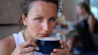 Dolu kafede cappuccino içen genç bir kadın, şehir hayatının ortasında huzurlu bir an yaşıyor, sakin bir yüz ifadesiyle aromatik içeceğin tadını çıkarıyor. Her gün kahve tüketimi kavramı.