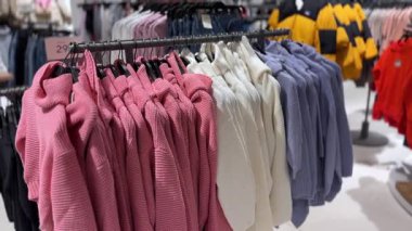 Sıcak pembe, beyaz ve mor kazaklar bir moda mağazasında bir elbise rafında asılı, müşterilerin daha soğuk mevsimler için sıcak ve şık bir seçenek sunmaya ve satın almaya hazır.