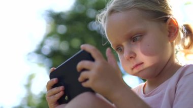 Genç sarışın anaokulu öğrencisi hareketli oyunlara dalmış, akıllı telefonlu çimlerin üzerinde oturuyor, güneşli bir yaz öğleden sonrası boyunca yeşil parklarla çevrili. Çocuklar ve aletler için kavram