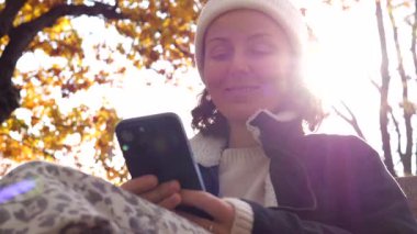 Güneş ışığı altın yaprakların arasından parlarken beyaz bir bere takan ve siyah ceketli bir kadın bir bankta oturuyor, akıllı telefonunu kullanırken gülümsüyor. İnternet 'ten bilgi tüketimi kavramı