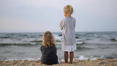 İki genç çocuk, muhtemelen kardeşler ya da arkadaşlar, kumlu bir sahilde durup, denizin engin engin genişliğine bakıyorlar, düşüncelerinde kayboluyorlar ve huzurlu atmosferin tadını çıkarıyorlar.