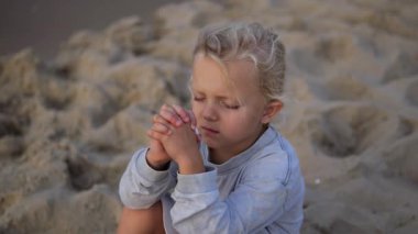 Sarışın çocuk, gözleri kapalı, elleri dua ederken kenetlenmiş, dingin plaj meditasyonu sırasında derin manevi anlar yaşıyor.