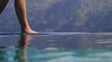 Sonsuz bir havuzun su yüzeyinde çıplak ayakla yürüyen ve arka planda bir dağ manzarası olan kadın bacakları özgürlük ve sükunet hissi yaratıyor.