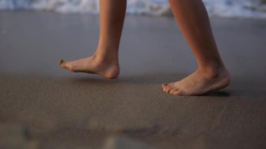 Çıplak ayaklı çocuk deniz kıyısında yürüyor, ayak parmaklarının arasındaki yumuşak dalgaların ve yumuşak kumların tadını çıkarıyor, huzurlu ve kaygısız bir yaz sahnesi yaratıyor.