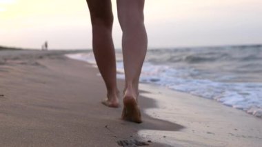 Kumlu deniz kıyısında çıplak ayakla yürüyen kadın, altın gün batımında okyanus dalgalarının altında sakin bir akşamın tadını çıkarırken yumuşak ayak izleri bırakıyor.