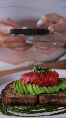 Dikey ekran, akıllı telefon kullanan kadın soslu domatesli avokado tostunun fotoğrafını çekiyor ve soslu ekmeği sosyal medyada paylaşmaya hazır.