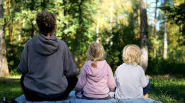 Yoga eğitmeni iki küçük çocuğa huzur dolu bir orman ortamında rehberlik ediyor, meditasyon ve nazik pozlarla farkındalığı ve refahı teşvik ediyor. Yoga konsepti ve çocuklarla meditasyon