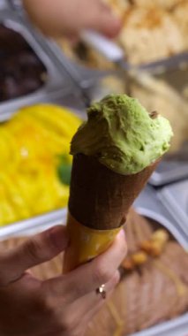 Dikey ekran, kremalı şam fıstıklı dondurmayı koniye batıran satıcı, arka planda gösterilen renkli gelato çeşitleri, canlandırıcı yaz tatlısını temsil ediyor.