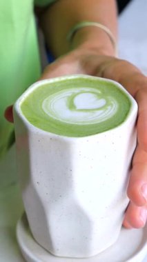 Dikey ekran, kadın kahveci elleri kalp şeklindeki latte sanatıyla bir fincan kibrit latte tutuyor, bardağın farklı açılarını ve perspektiflerini gösteriyor.