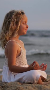 Dikey ekran, dingin genç kız yoga yapıyor, kumlu sahilde sakin okyanus dalgalarıyla huzur içinde meditasyon yapıyor.