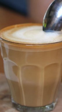 Dikey ekran, profesyonel barista kremalı köpük yaratıyor ve kırsal ahşap masada şeffaf bir bardak kahveye dikkatlice köpüklü tabaka ekliyor.