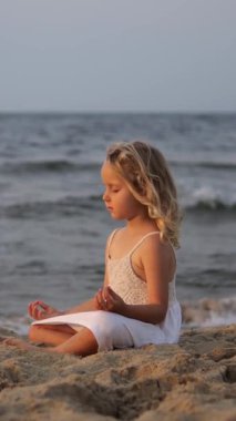 Dikey ekran, kumlu sahilde meditasyon yapan sarışın çocuk, altın gün batımında hafif okyanus dalgalarını dinlerken iç huzur ve sükunet arıyor.
