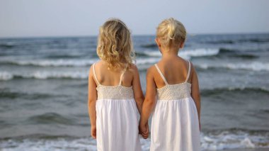 Sarışın kız kardeşler el ele tutuşmuş el ele tutuşmuş saf beyaz elbiseler giyip altın saat boyunca engin deniz manzaralarına bakıyorlar çocukluk masumiyetini ve aile bağlarını somutlaştırıyorlar.