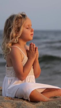 Dikey ekran, beyaz elbiseli barışçıl sarışın kız gün batımında kumsalda meditasyon yapıyor, elleri duada birleşiyor, dingin ve ruhani bir atmosfer yaratıyor.