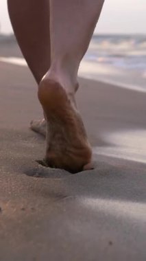 Dikey ekran, ıslak kumda çıplak ayakla yürüyen kadın bacakları, altın gün batımında sahil kenarında ayak izleri bırakarak, huzurlu kıyı keyfi ve yaz tatili atmosferini somutlaştırıyor.