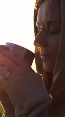 Dikey ekran, arka planda kapüşonlu sweatshirt giyen bir kadın nefes kesen bir gün doğumunda sıcak bir içeceğin keyfini çıkarıyor, huzurlu ve huzurlu bir atmosfer yaratıyor.