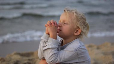 Sarışın, genç bir kız, gözleri kapalı, sakin bir şekilde el ele tutuşuyor, sakin okyanus dalgalarının yanında, kumlu sahil kıyısında altın gün batımında.