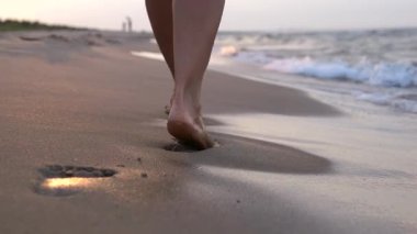 Kumlu sahil boyunca çıplak ayakla yürüyen dişi bacaklar ıslak kumda desenler oluştururken yaz tatilinde altın gün batımı ışığı ve sakin okyanus manzarasının tadını çıkarıyor.