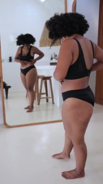 Dikey ekran, güzel ve iri yarı siyah kadın siyah iç çamaşırlarıyla vücudunun pozitifliğini kucaklıyor ve ev ortamında kendini sevdiriyor.