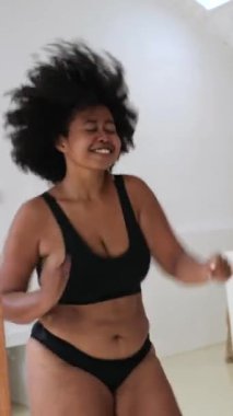 Dikey ekran, kıvrımlı model siyah iç çamaşırlarıyla güven içinde dans ediyor, kıvırcık saçlarını parlak beyaz odada enerjik hareketlerle sallıyor.