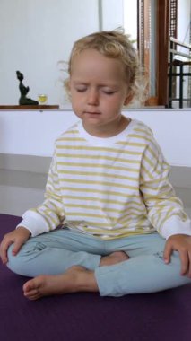 Dikey ekran, günlük kıyafetler giyen sarışın anaokulu çocuğu mor yoga minderinin üzerinde lotus pozisyonunda oturuyor, dikkatle ve kapalı gözlerle meditasyon yapıyor.