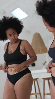 Dikey ekran, güzel ve iri yarı siyah iç çamaşırlı Afro-Amerikan kadın göbeğini kontrol ediyor ve bir evin içindeki aynanın önünde karnını çimdikliyor.