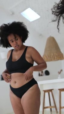 Dikey ekran, güzel ve iri yarı siyah iç çamaşırlı Afro-Amerikan kadın vücudunun yağını ölçüyor, karnını ve kolunu çimdikliyor, görünüşü ve kilosu hakkında endişe duyuyor.