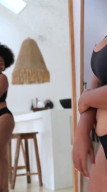 Dikey ekran, kendine güven artı beden modeli kendini beğenme ve vücut kabullenmeyi gösteriyor, minimalist bir banyo ortamında ayna ve ahşap bir taburede siyah iç çamaşırlarıyla poz veriyor.