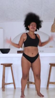 Dikey ekran, afro saçlı, siyah iç çamaşırı giyen, modern bir mutfakta neşe içinde dans eden, özgürlük ve kendini seven genç bir kadın.