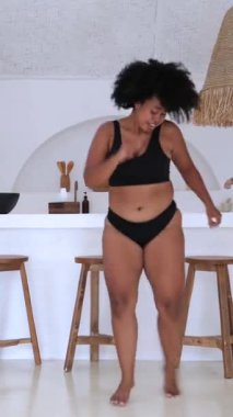 Dikey ekran, güzel, vücut pozitifliği, artı uzun saçlı zenci kadın neşeyle dans ediyor ve minimalist bir mutfakta iç çamaşırıyla özgürce kendini ifade ediyor.