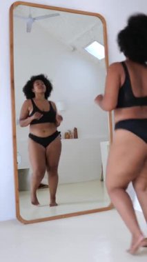 Dikey ekran, güzel, kıvrımlı, uzun saçlı, siyah iç çamaşırı giyen, modern, minimalist bir iç mekanda aynanın önünde dans eden bir kadın.