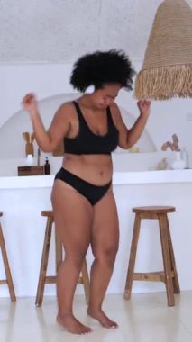 Dikey ekran, kıvırcık saçlı, siyah iç çamaşırlı, beyaz kulaklıklı güzel bir kadın. Minimum beyaz bir odada ahşap tabureler ve bir tezgahla enerjik bir şekilde dans ediyor.