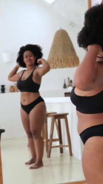 Dikey ekran, güzel ve iri yarı Afro-Amerikan kadın siyah iç çamaşırı giyiyor. Vücut şeklini evde aynanın önünde kontrol ediyor. Kıvrımlarıyla rahat ve kendinden emin.
