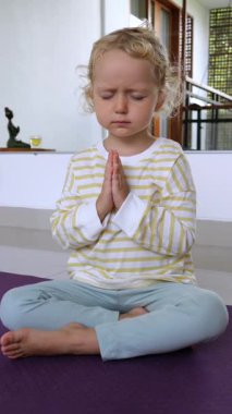 Dikey ekran, gözleri kapalı barışçıl sarışın çocuk, mor yoga minderinde lotus pozisyonunda oturuyor, dua pozunda birlikte meditasyon yapıyor.
