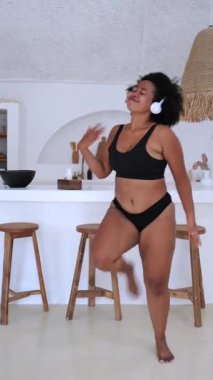 Dikey ekran, Afro-Amerikan kadın siyah kablosuz kulaklıklar ve iç çamaşırlarıyla neşeyle dans ediyor, modern beyaz mutfak alanında kendine güven yayıyor.