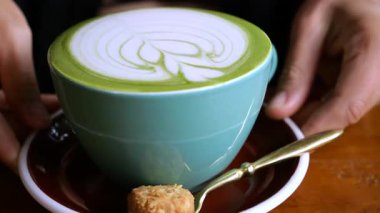 Barista, üzerinde altın bir kaşık ve gevrek bir bisküvi bulunan, latte sanatıyla süslenmiş turkuaz bir latte servis ediyor.