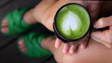 Ahşap verandada dinlenen, yeşil terlik giyen, elinde kalp şeklinde köpük sanatı olan kibrit latte tutan, tatilde huzur ve refah dolu bir anın tadını çıkaran bir kadın.