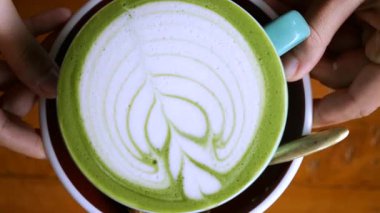 Yetenekli barista karışık köpüklü latte sunar, beşikteki turkuaz kupa ve tabağı yıpranmış ahşap yüzey üzerinde, mutfak işçiliği sergilenir.