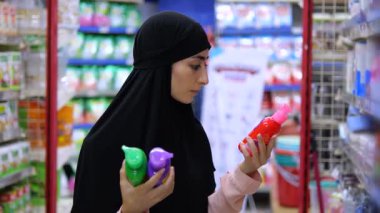 Siyah tesettürlü genç Müslüman kadın süpermarket raflarındaki temizlik ürünlerini karşılaştırıyor, farklı deterjan şişelerini dikkatlice analiz ediyor ve satın alma kararı veriyor.