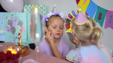 Sarışın kız, renkli dekorasyonlar ve şenlikli atmosferle çevrili bir doğum günü partisinde arkadaşlarının yüzüne dikkatlice makyaj yapıyor.