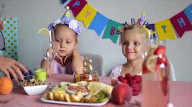 İkiz kardeşler süslü şapkalar takıp renkli sağlıklı atıştırmalıklar ve içeceklerle süslü masada oturuyorlar. Doğum günü partisinin başlamasını beklerken beklenti ve heyecan gösteriyorlar.
