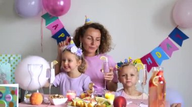 Anne ve iki kızıyla beşinci doğum gününü kutlayan aile renkli balonların, hediyelerin, hediyelerin arasında parti şapkaları takıp evde neşeli bir ortam yaratıyor.