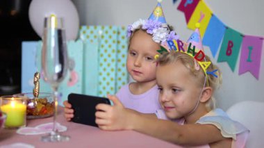 Doğum günü şapkaları ve parti elbiseleri giyen iki genç kız kardeş, doğum günü partilerinde hediye, balon ve hediyelerle çevrili akıllı telefonlarıyla selfie çekiyorlar.