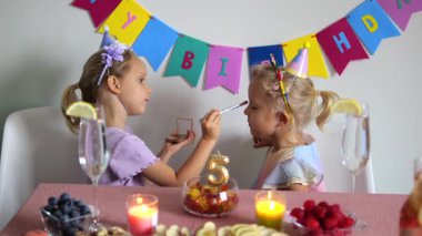 İki genç kız kardeş beşinci doğum gününü mum, meyve, fındık ve içeceklerle kutlarken, bir kız kardeş diğerine şakayla makyaj yapıyor.