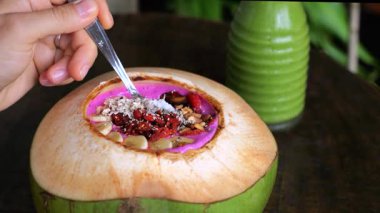 Elleri kaşıkla taze hindistan cevizi kabuğunun içinde yeşil meyve suyu şişesinin yanında sunulan granola, tohum ve hindistan cevizi taneleriyle dolu canlı pembe bir smoothie kasesini kaldırıyor.